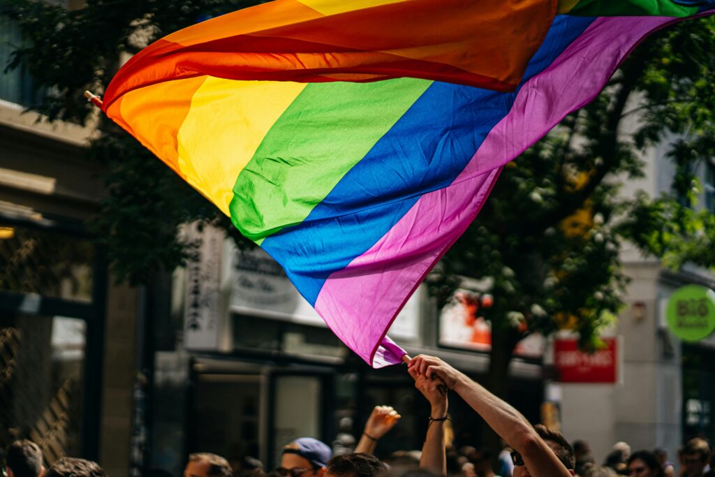 picture of a pride flag that is being held up by members of a crowd at a pride parade. The pride flag is very large.